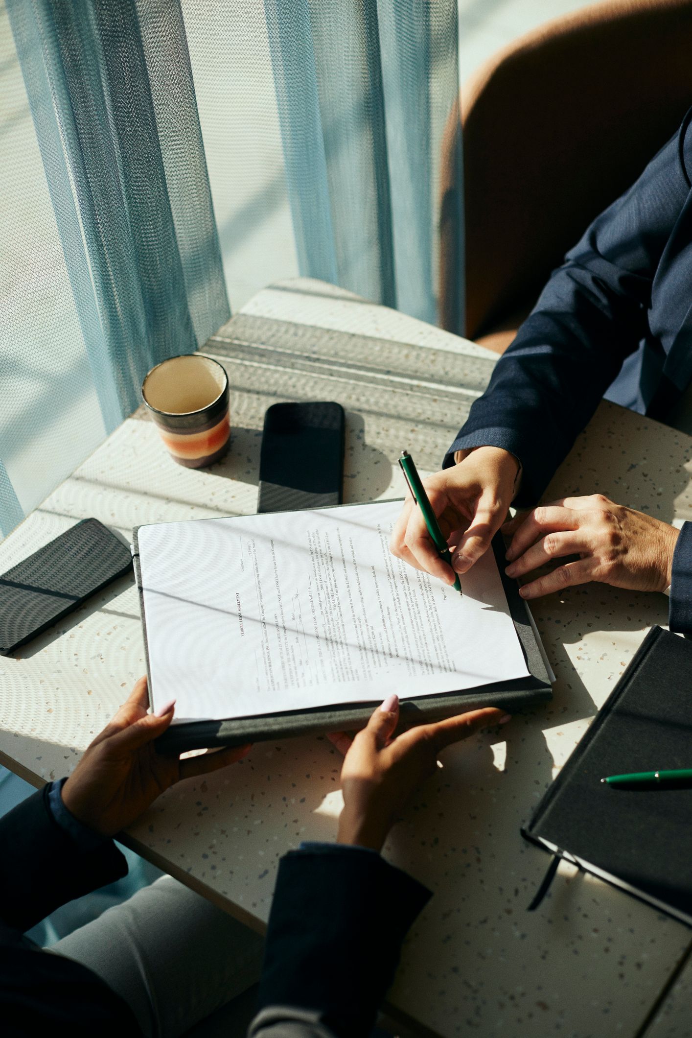 Two people at a table, one signing a document. A phone, coffee, and notebook are on the table.