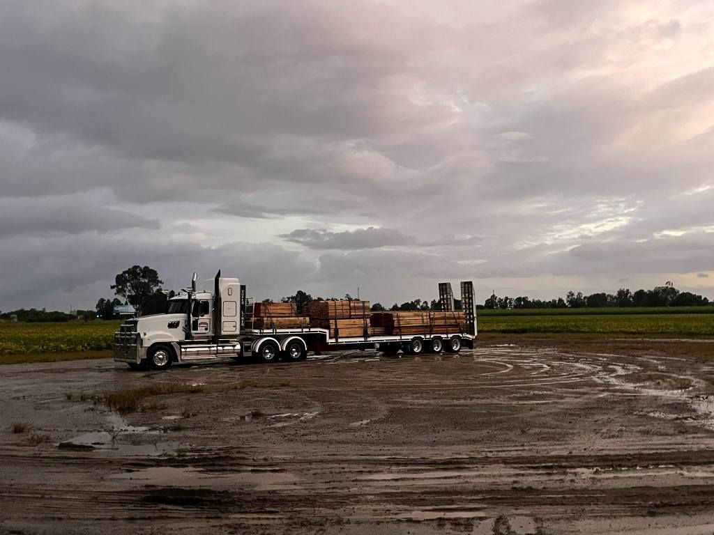 White Truck Hauling an Orange Logging Trailer Across a Muddy Field Under a Cloudy Sky — Rainbow and Sons Heavy Haulage in Coffs Harbour, NSW