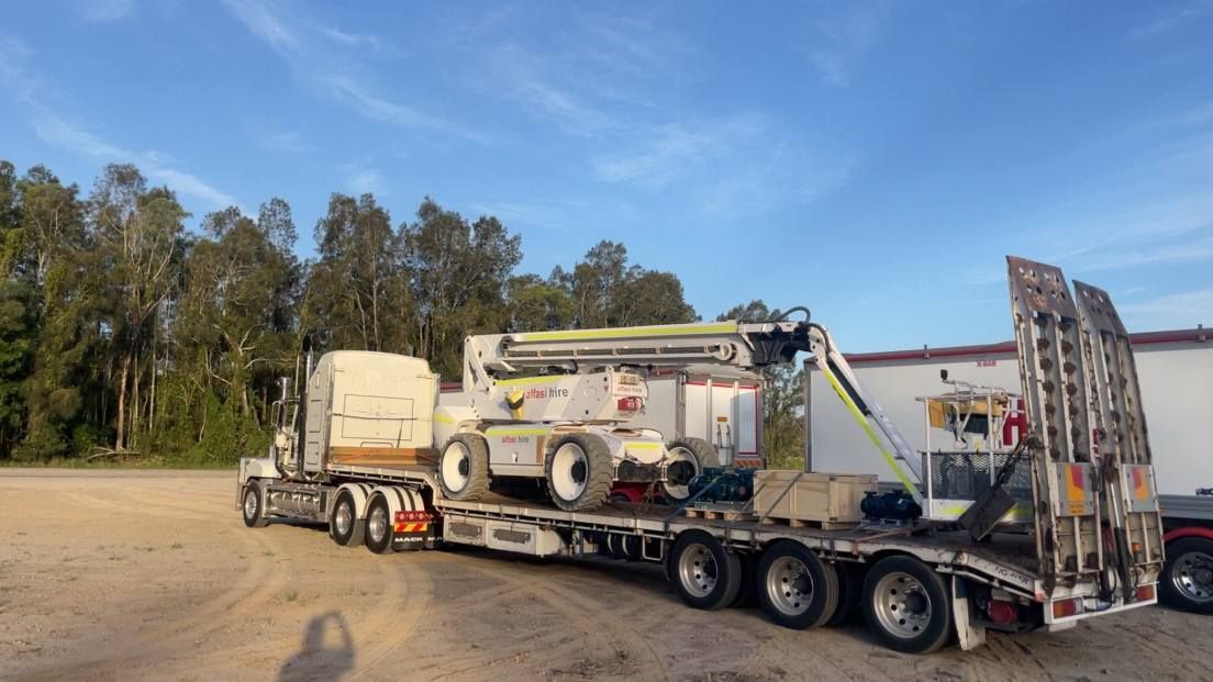 Flatbed Truck Hauling a Large White Industrial Machine in a Dusty Lot Under a Blue Sky — Rainbow and Sons Heavy Haulage in Coffs Harbour, NSW