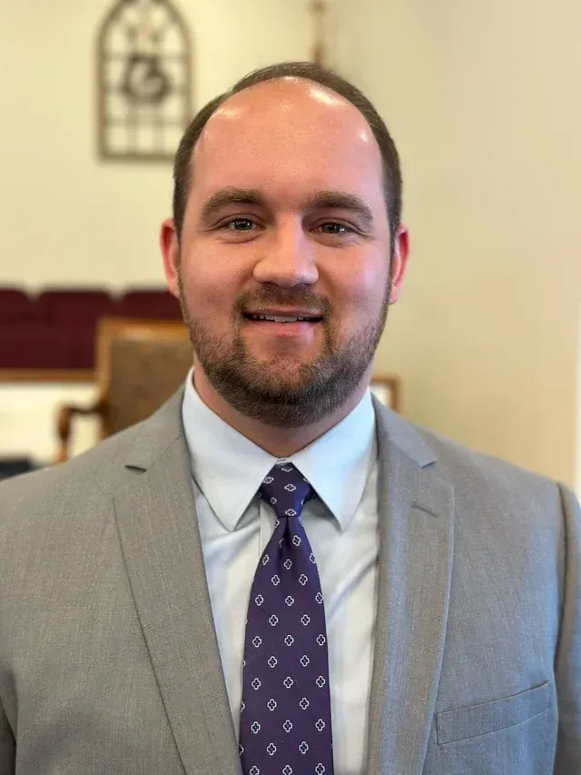 Man in gray suit with a purple patterned tie, smiling. Background includes a church interior.