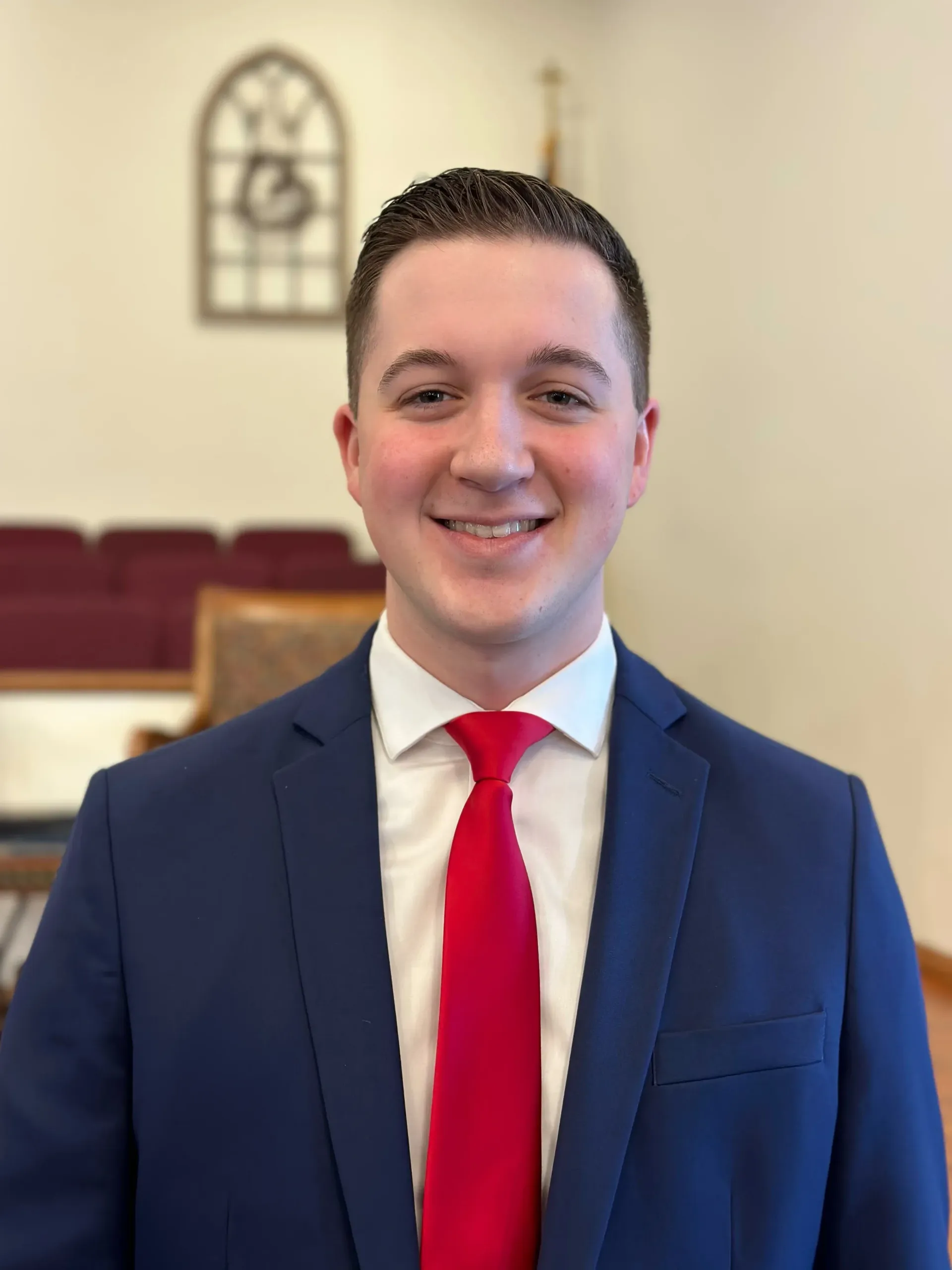 Man in a navy blue suit and red tie smiles in a church setting.