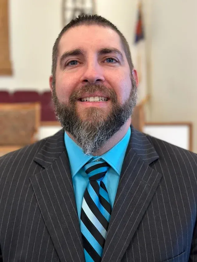 Man in dark pinstripe suit, blue shirt, and striped tie smiles. Blurred interior background.