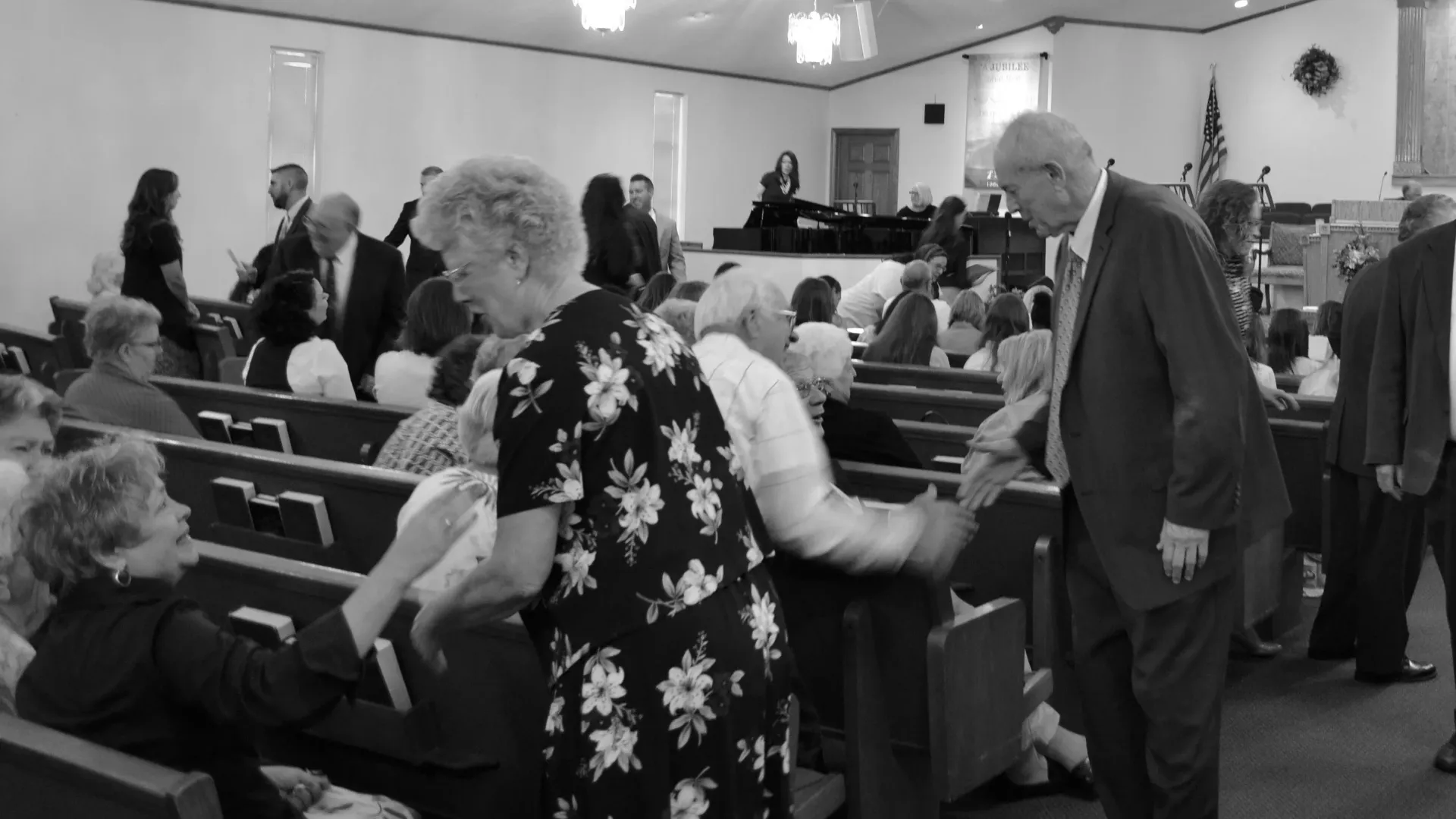 People in a church, some seated in pews, others greeting each other. Black and white photo.