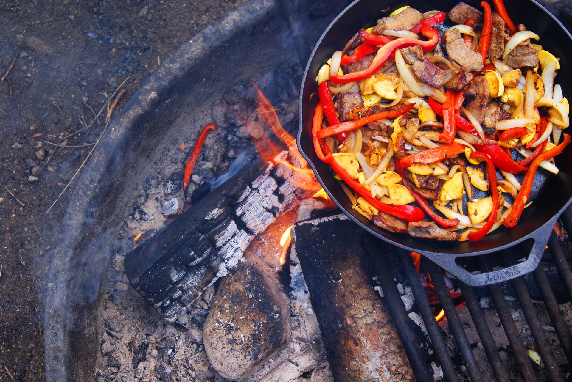 Cast iron skillet with food (steak, peppers, onions) cooking over a campfire.