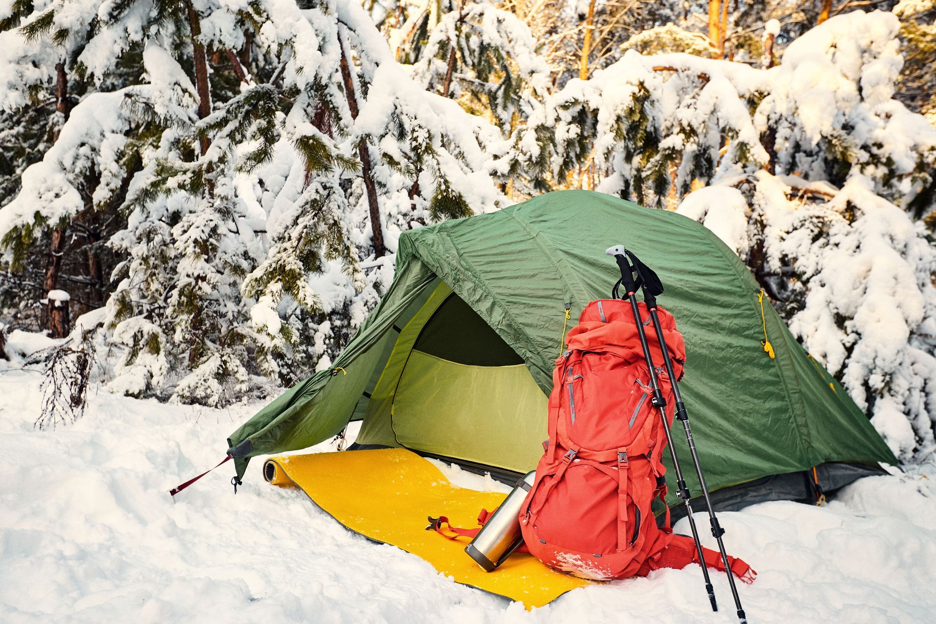 Green tent, red backpack, and hiking poles in a snowy forest. Yellow mat on snow.