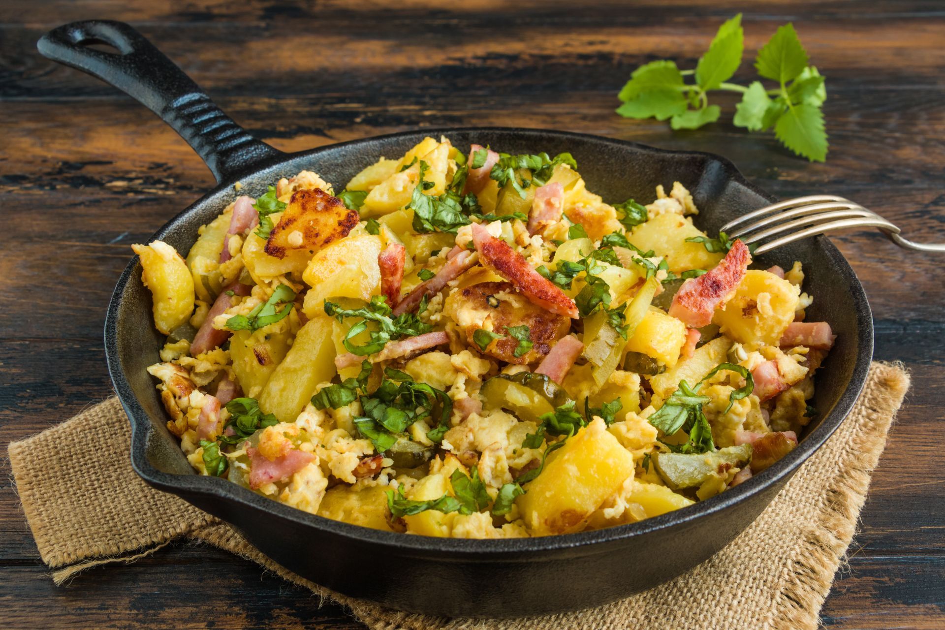 Potato hash with bacon and herbs in a black skillet on a burlap cloth, wooden table.