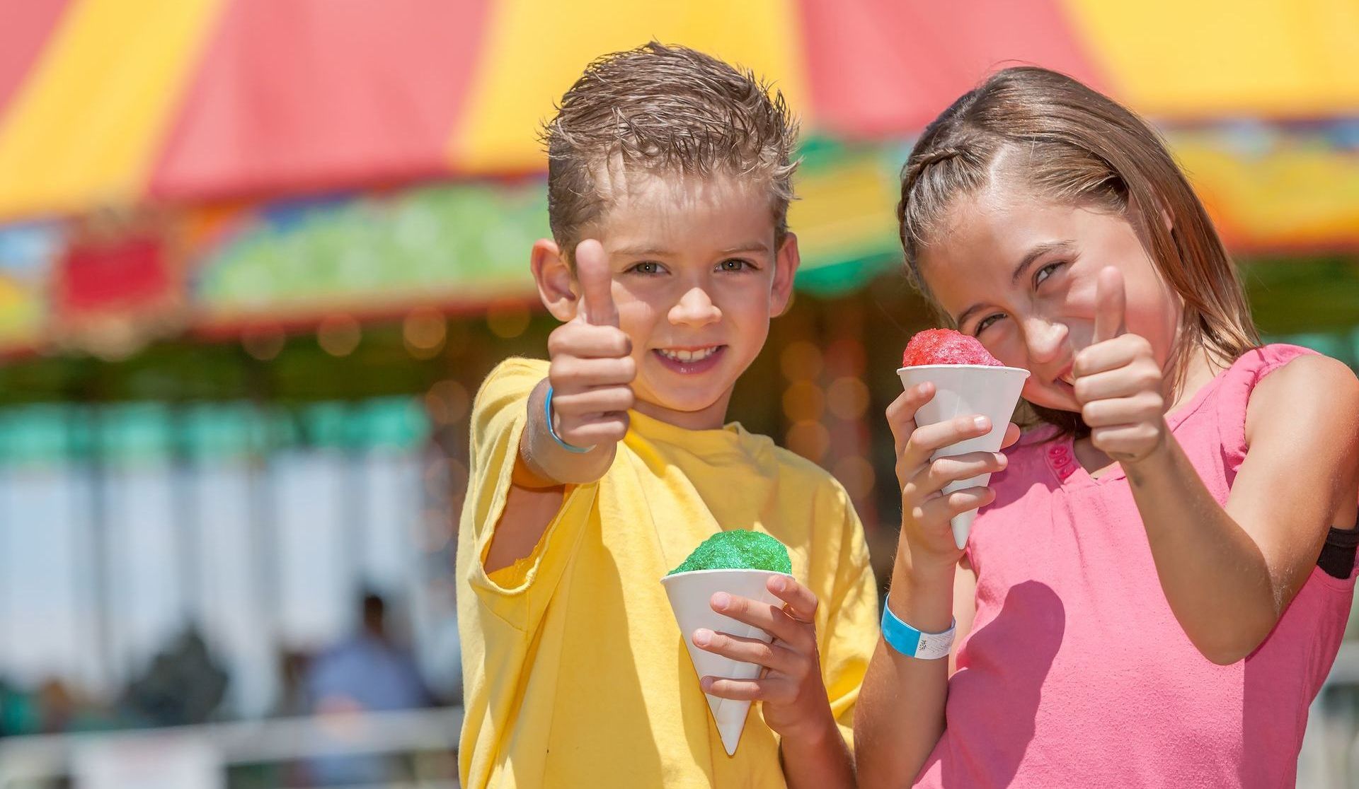 A boy and a girl are eating shaved ice at a carnival and giving a thumbs up.