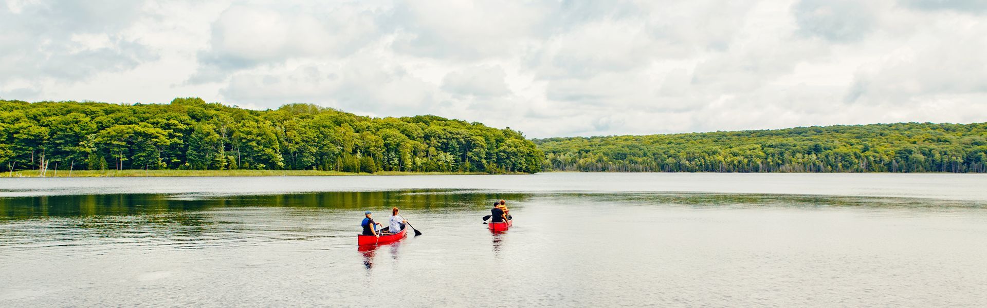 Two canoes with people on a calm lake, surrounded by green trees under a cloudy sky.