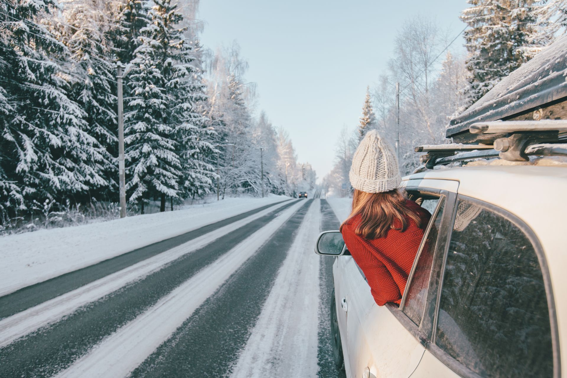 Woman leans out car window on snow-covered road lined by trees.