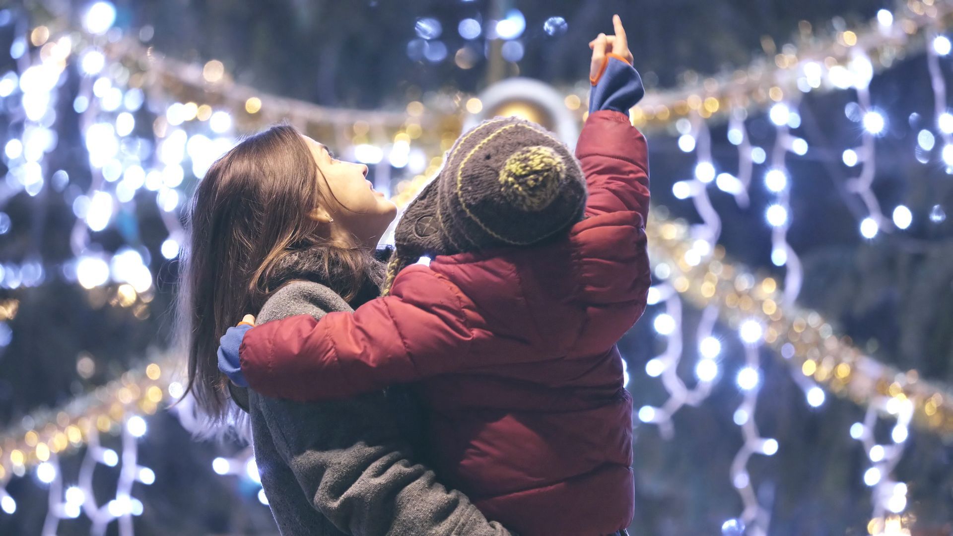 Woman holding a child, both looking up at a lit Christmas tree.