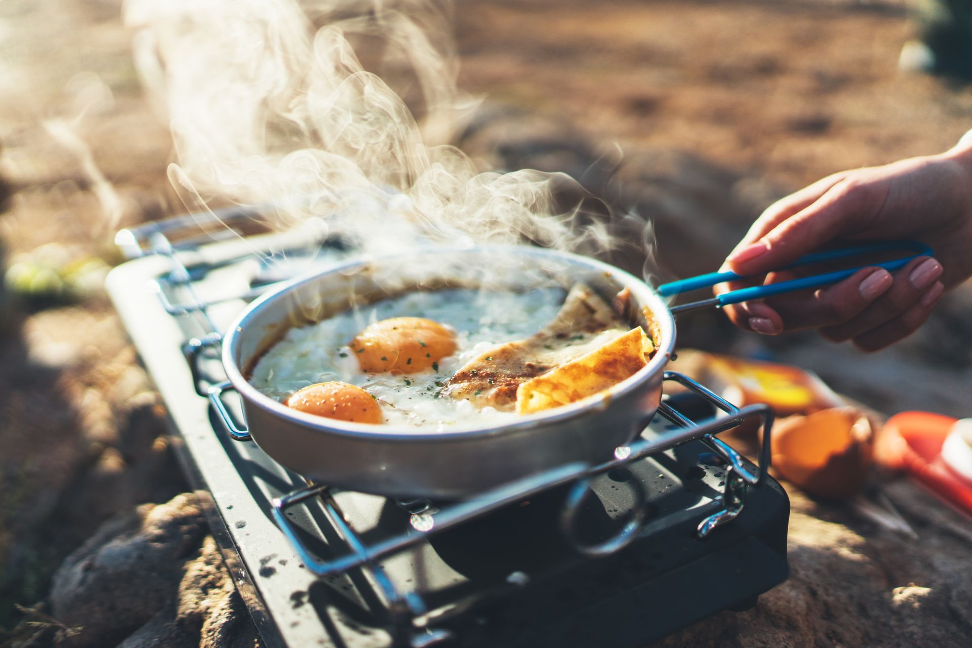 Eggs cooking in a pan over a portable camping stove; hand holding the handle.