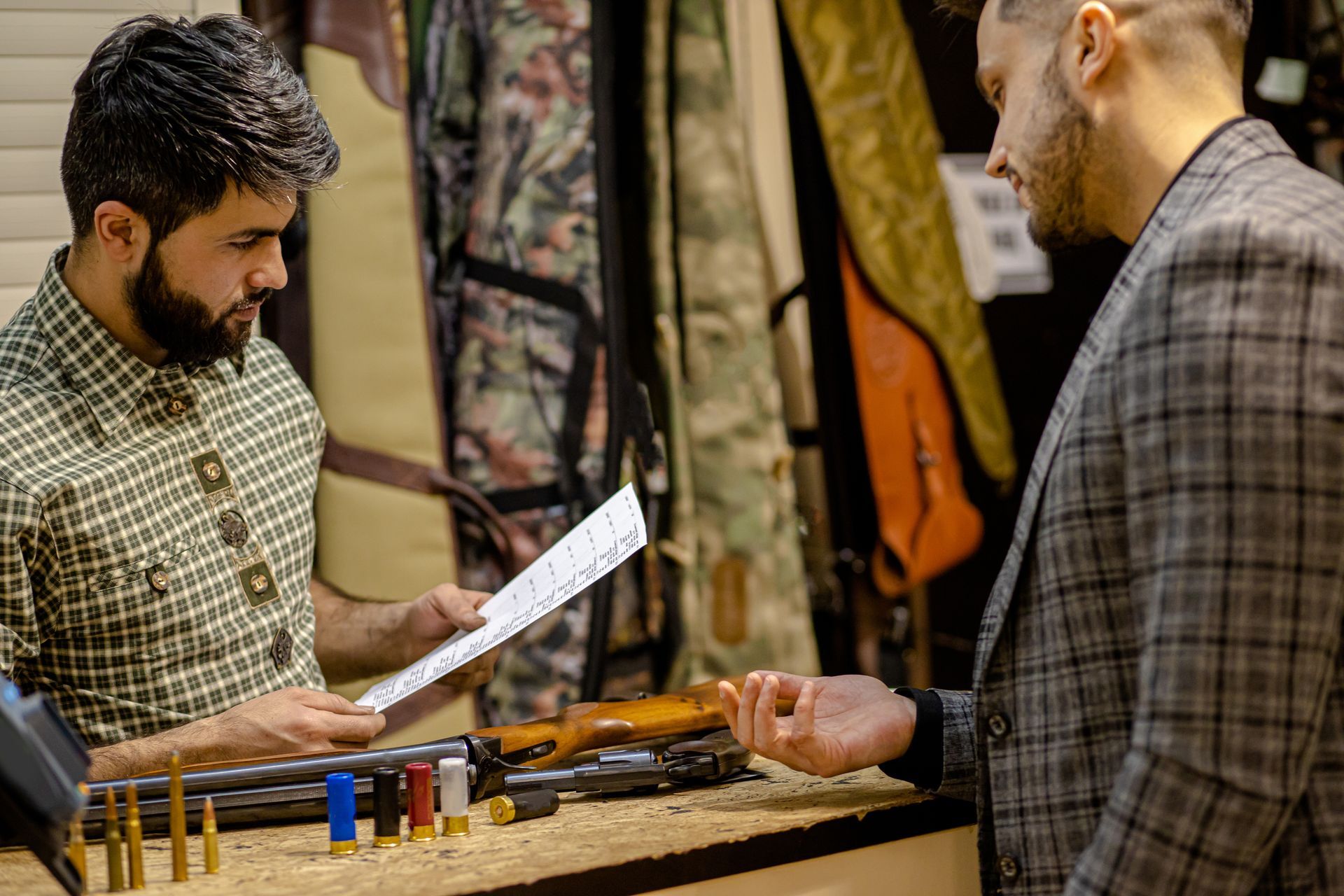 Two men examining paperwork and a shotgun at a store counter with ammunition and hunting gear.