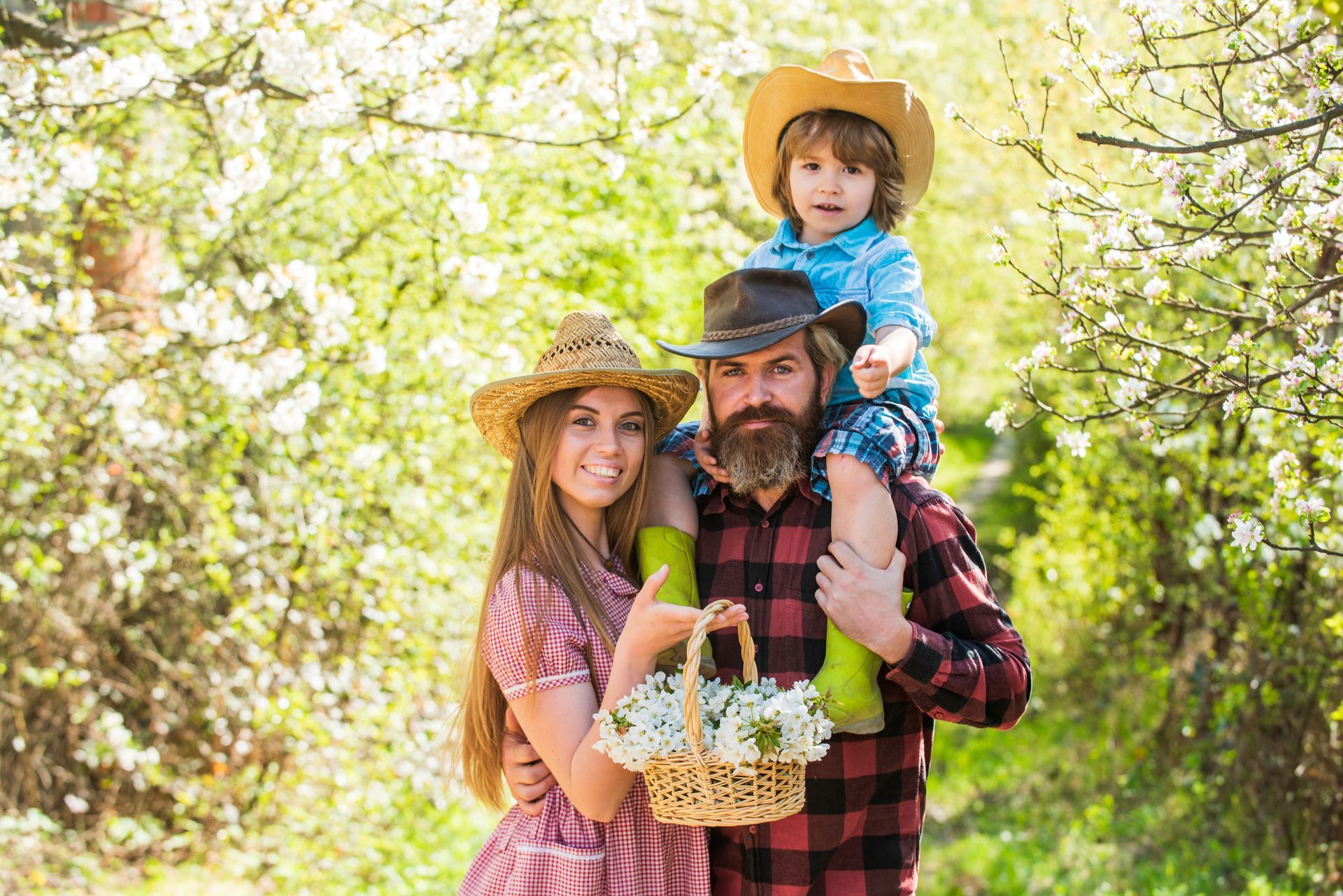 Family with a child on the father's shoulders, holding a basket of flowers, outdoors in a blooming orchard.