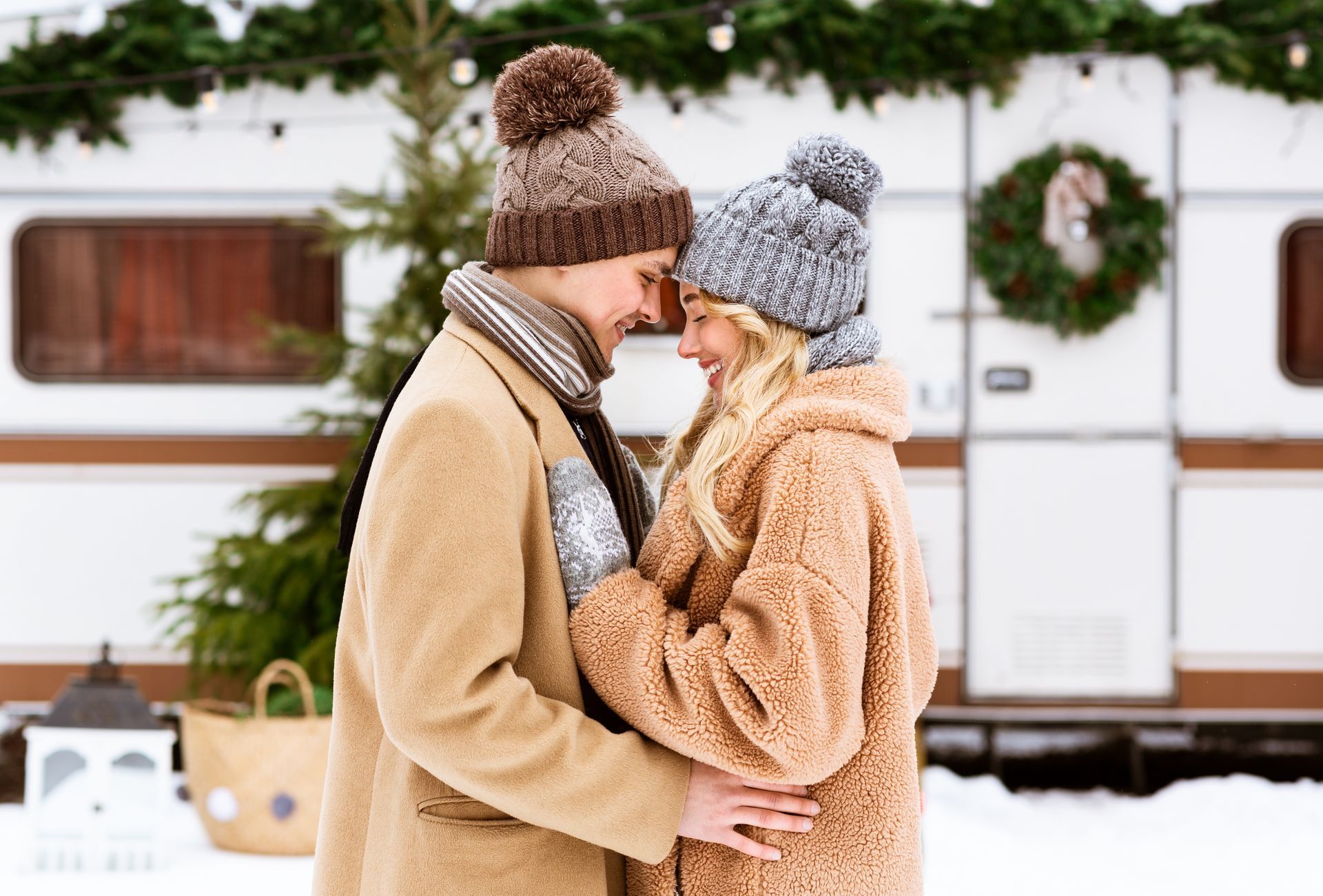 Couple in winter hats embrace in front of a camper, snowy scene.