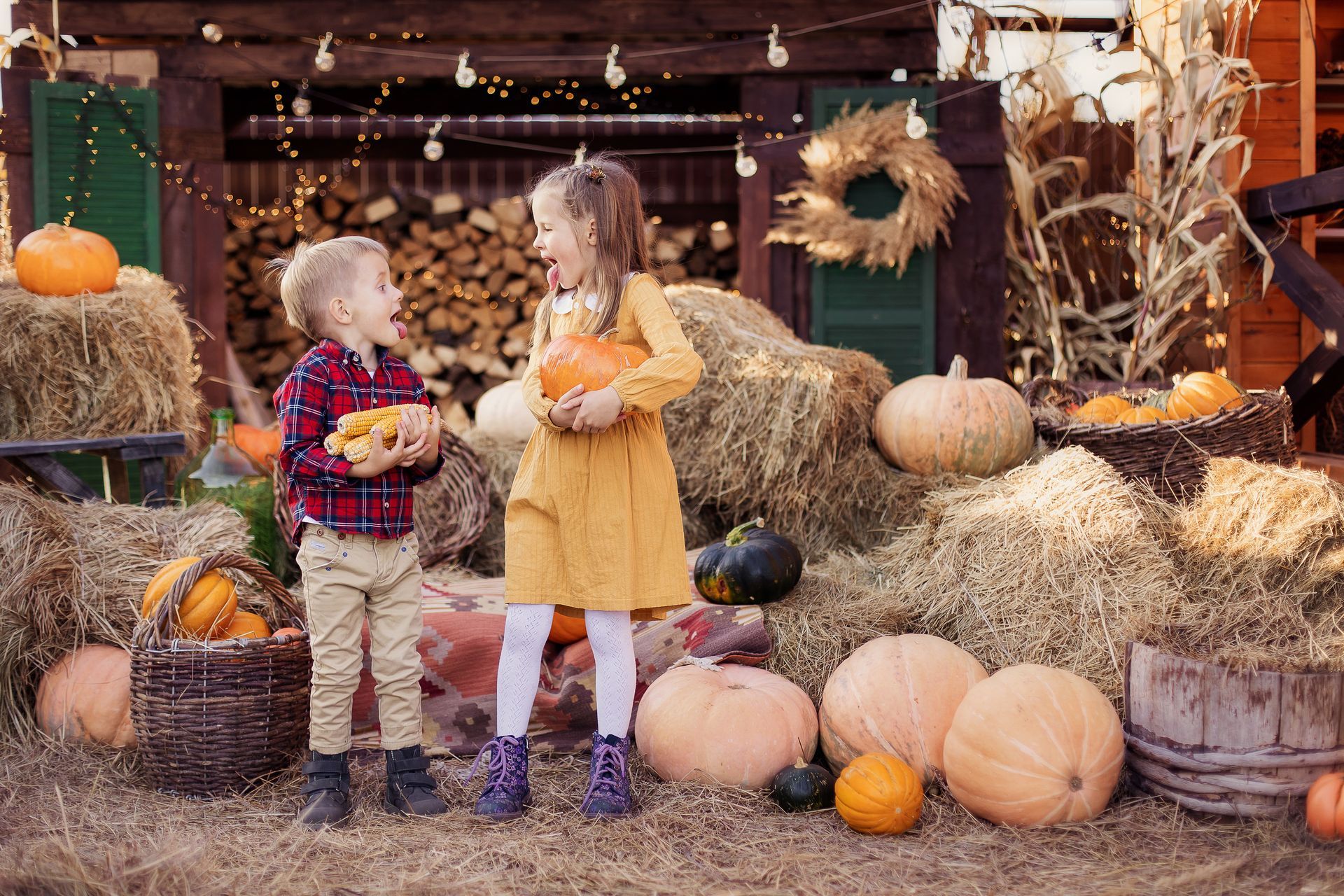 Boy and girl with pumpkins in a fall setting; straw bales, pumpkins, and decorative lights.