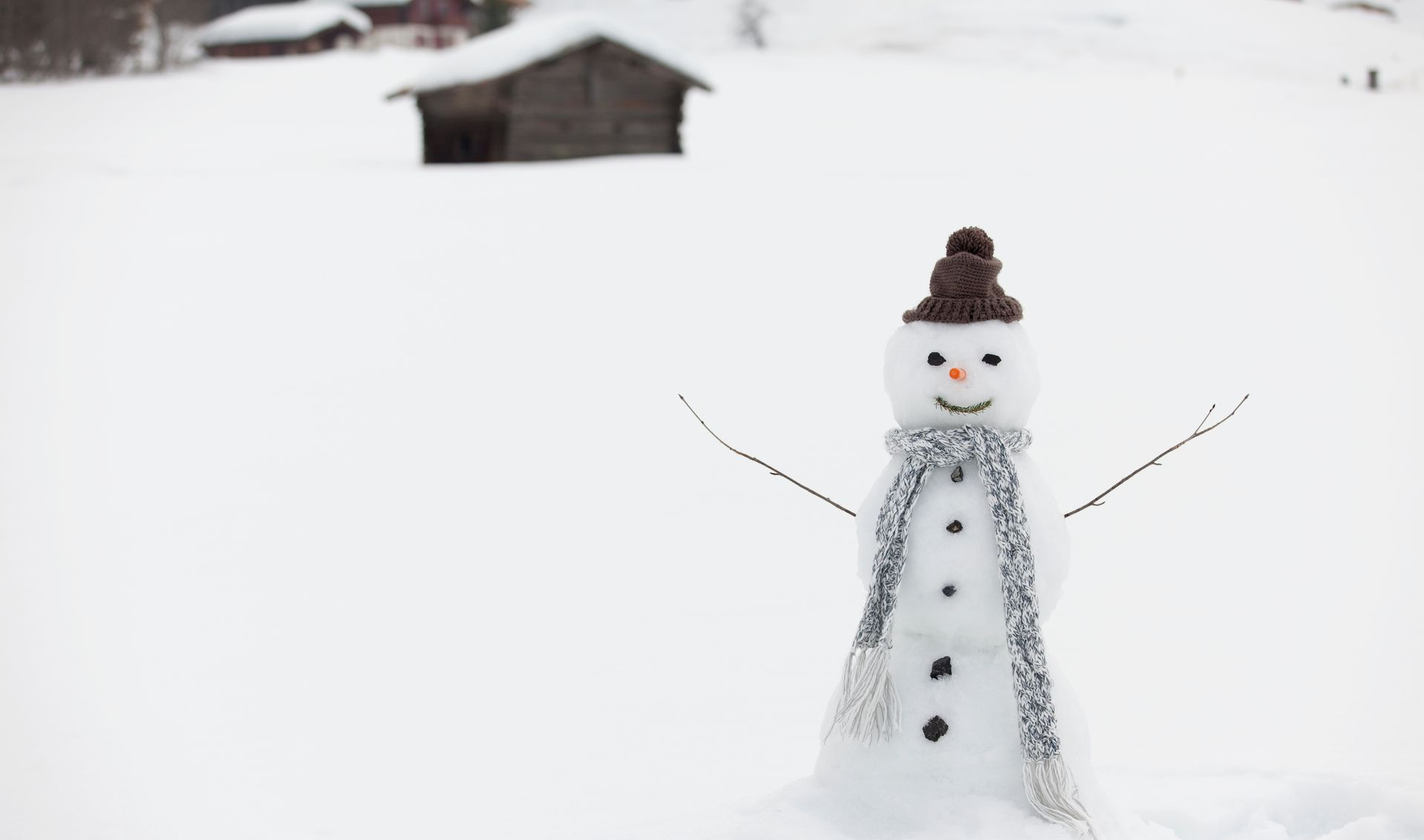 Snowman in a snowy field with a knitted hat and scarf, arms outstretched. Two wooden buildings in the distance.