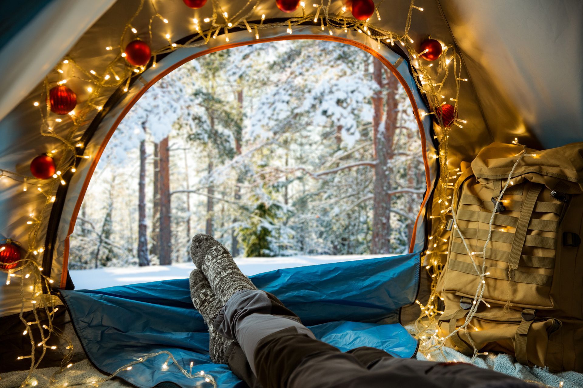 Person inside decorated tent, feet up, looking at snow-covered forest.