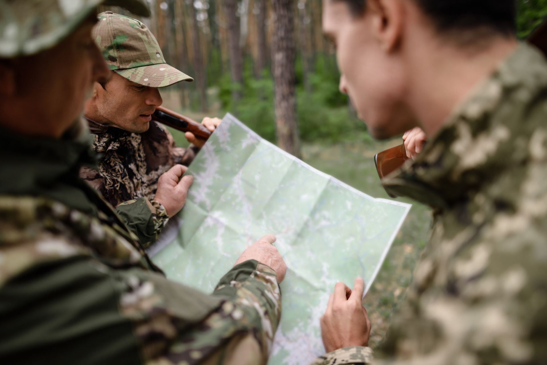Three people in camouflage looking at a map in a forest, pointing and discussing a route.