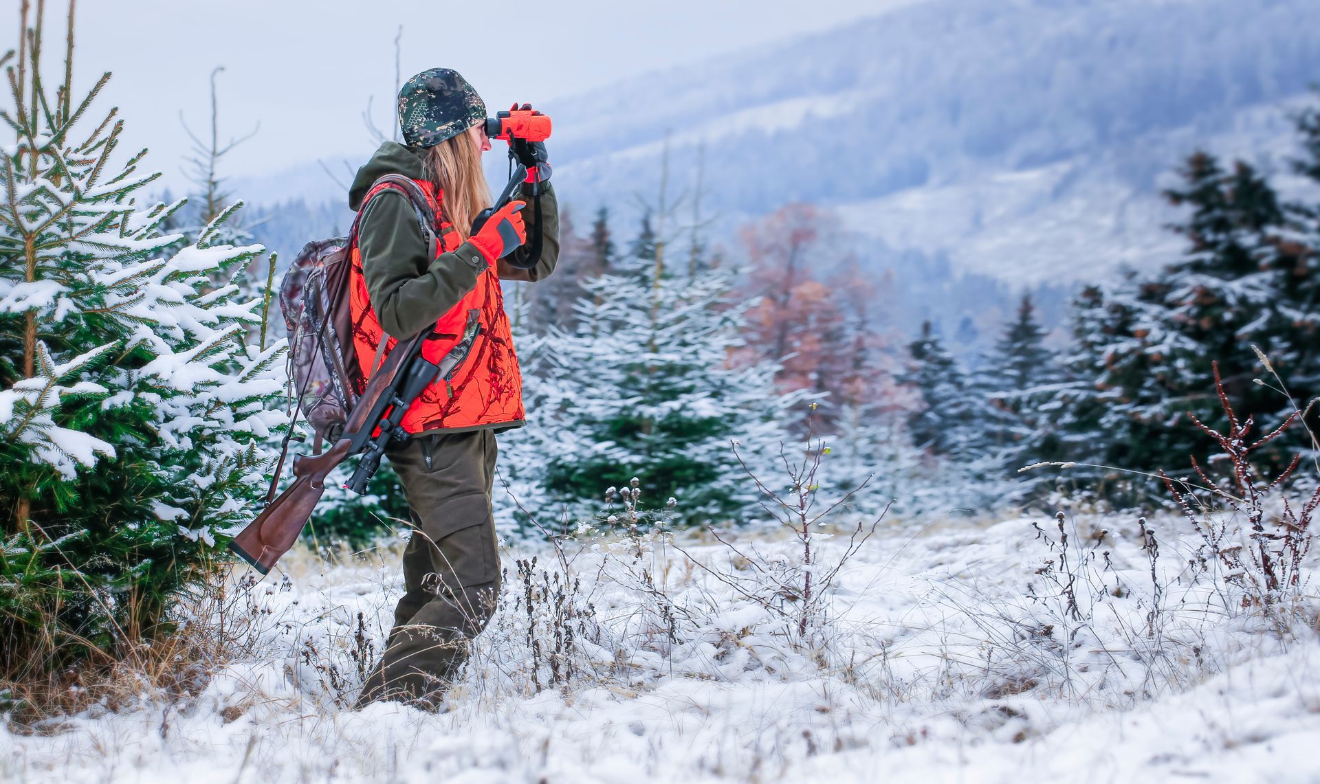 Hunter in orange vest, looking through binoculars in snowy forest with a rifle.