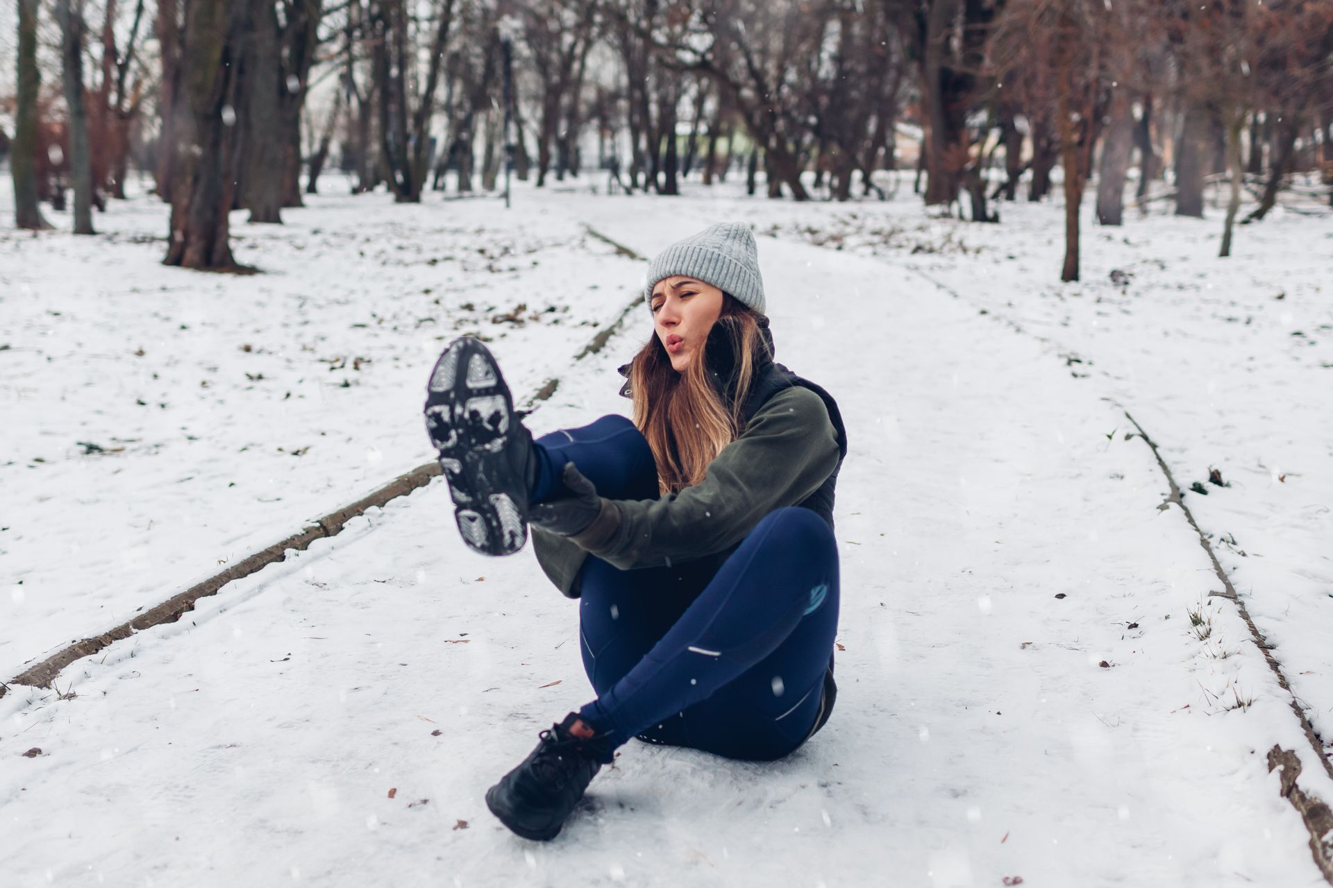 Woman sitting on snowy path, looking at her shoe, appears to have fallen.