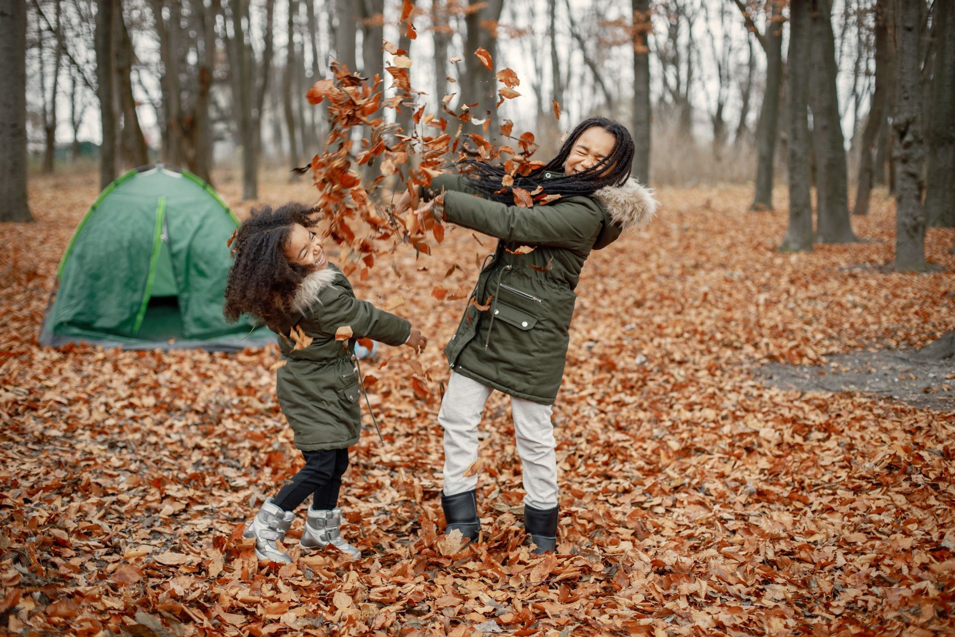 Two people in green coats throwing leaves in a forest with a tent.