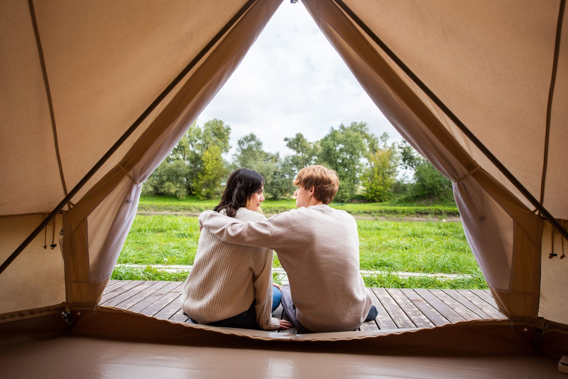 Couple sits together looking out from inside a tent, overlooking a grassy field with trees under cloudy skies.