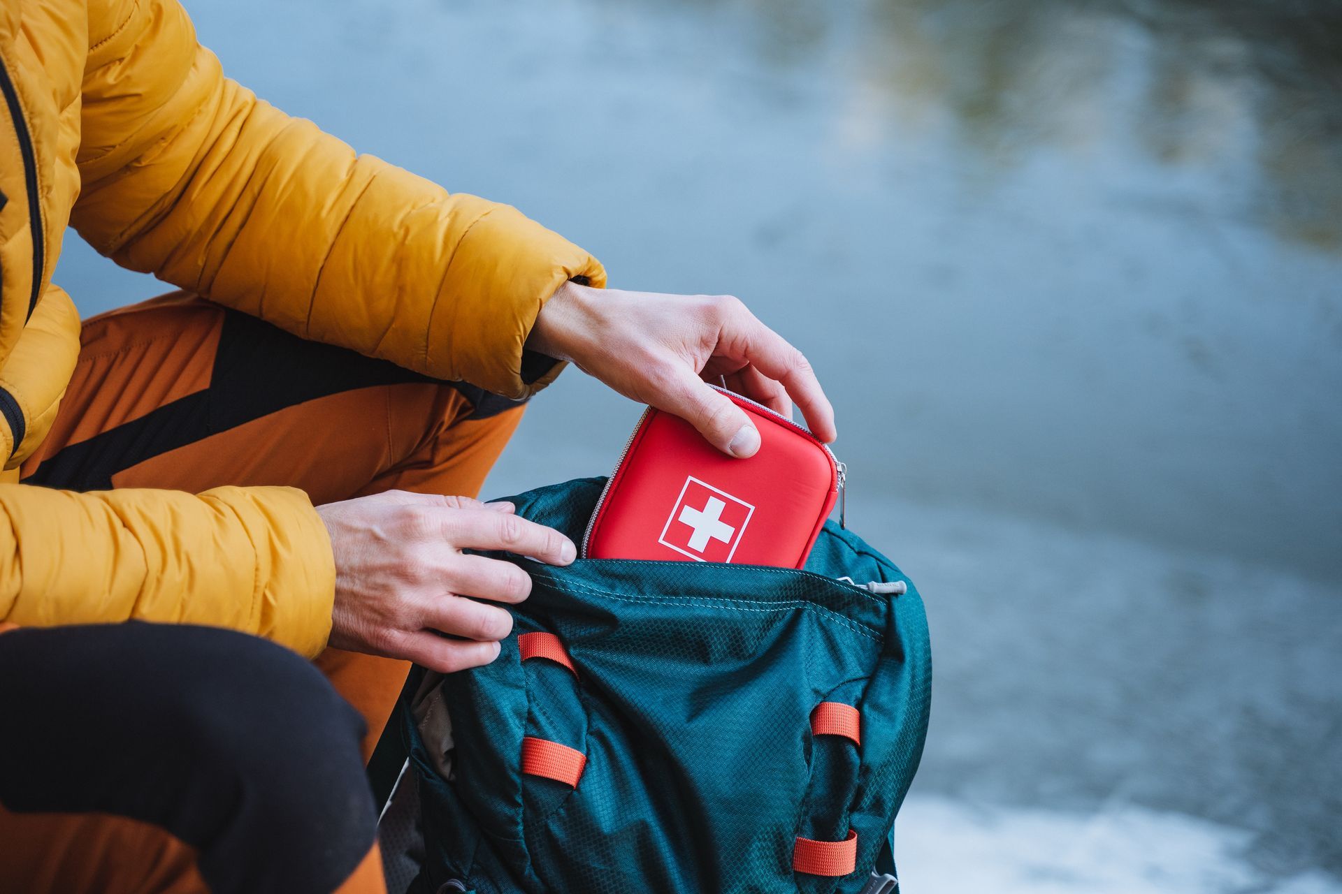 Person in yellow jacket placing a red first aid kit with a white cross into a teal backpack.