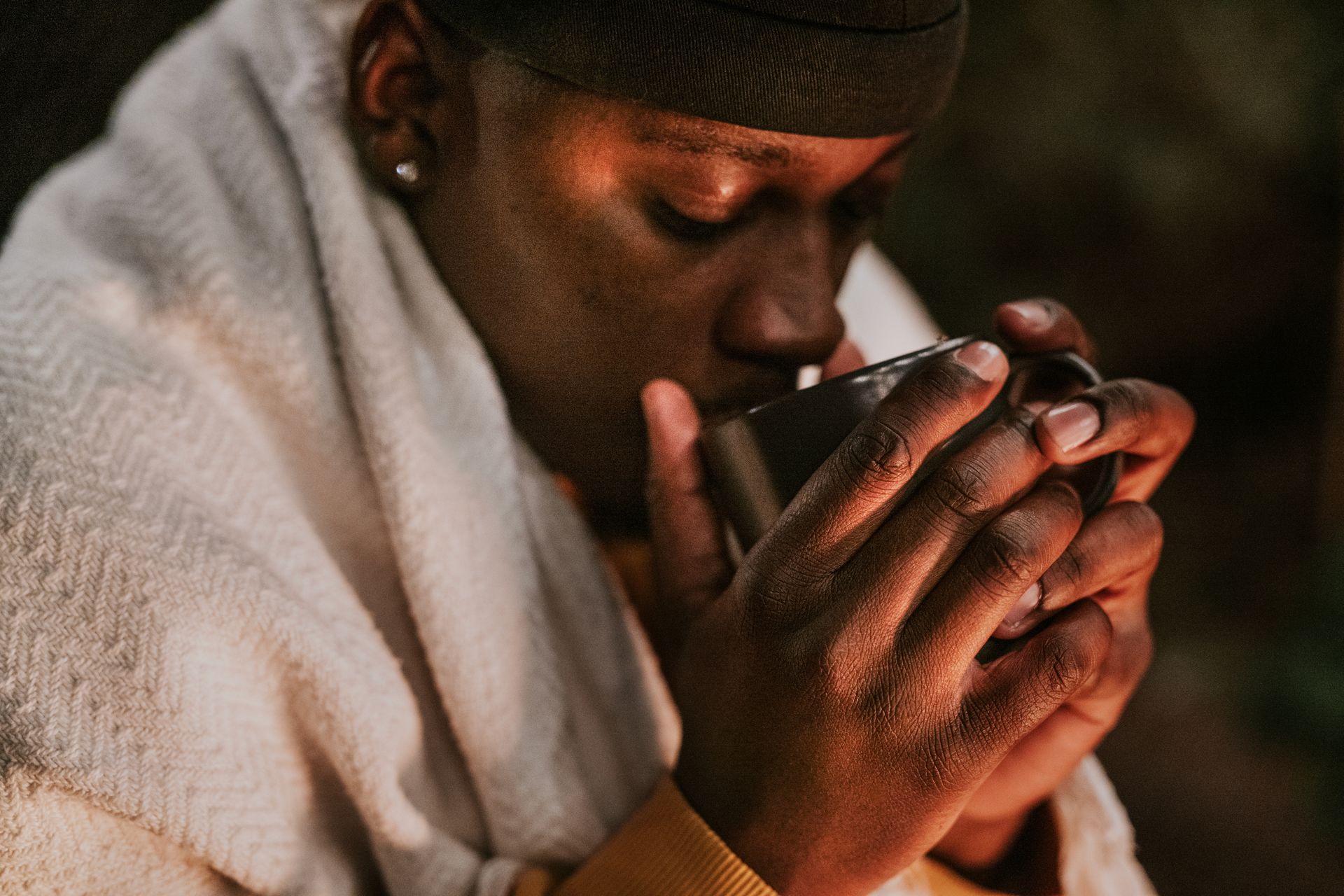 Woman wrapped in blanket, sipping from a black mug to keep warm.