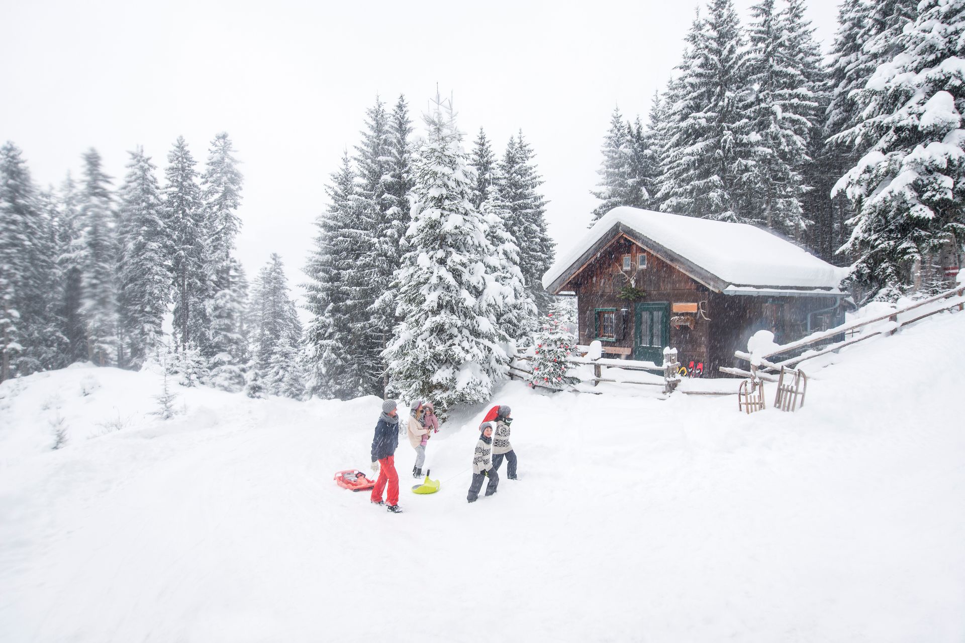 Snowy scene: Children near a cabin prepare to sled. Snow-covered trees and ground.