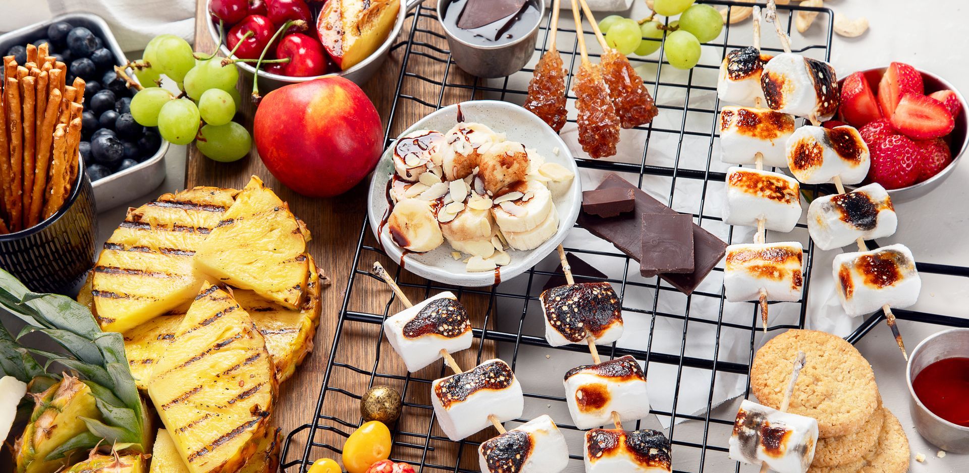 A table spread with various foods: fruit, grilled pineapple, marshmallows, bananas, chocolate, and dipping sauces.