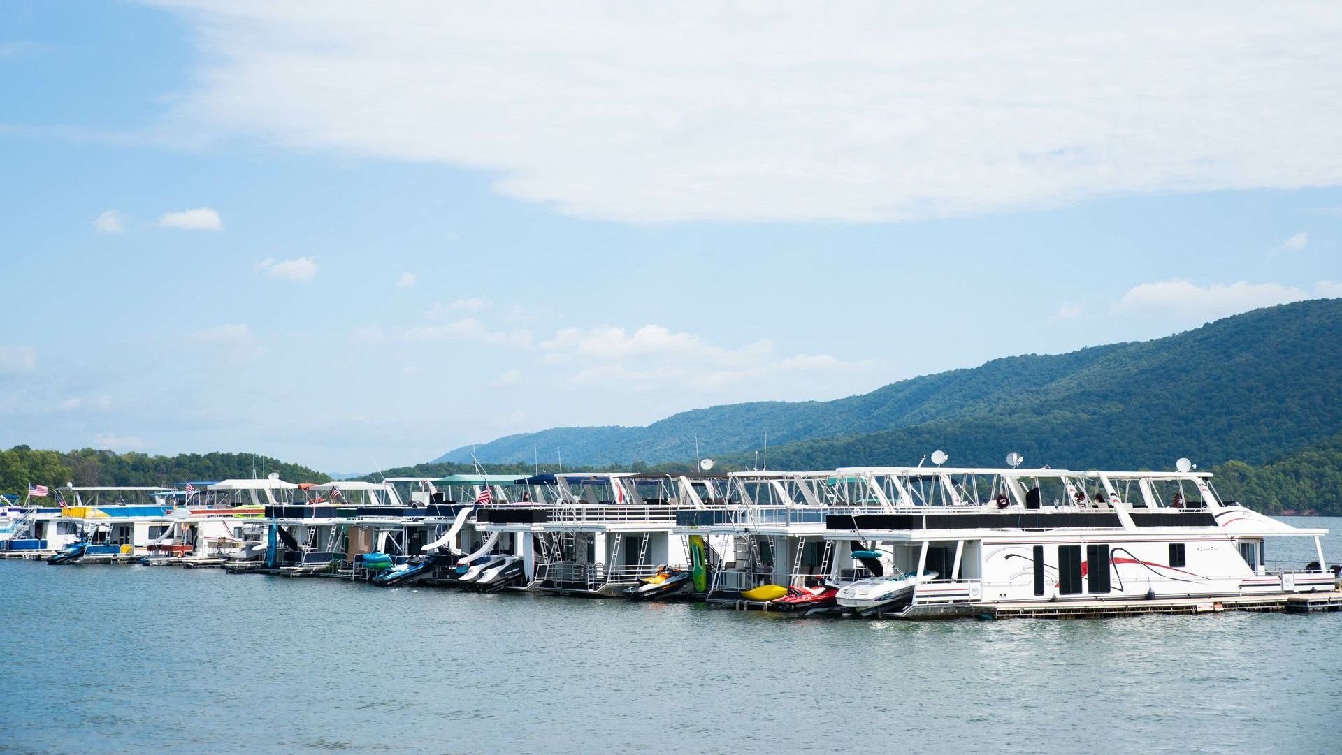 A row of boats are docked on a lake with mountains in the background.