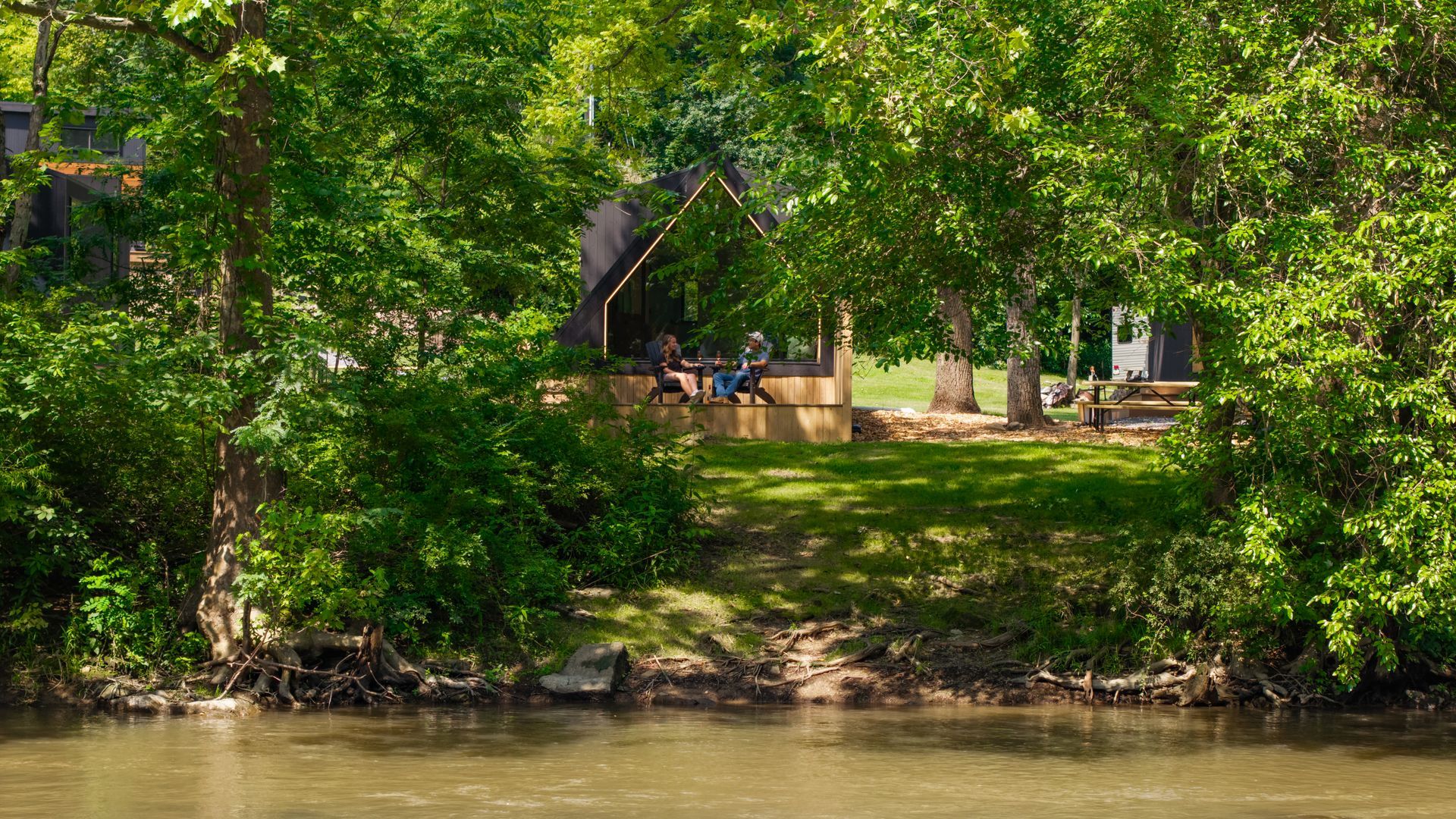 Two people sit on a wooden deck inside an A-frame structure overlooking a river surrounded by lush green trees.