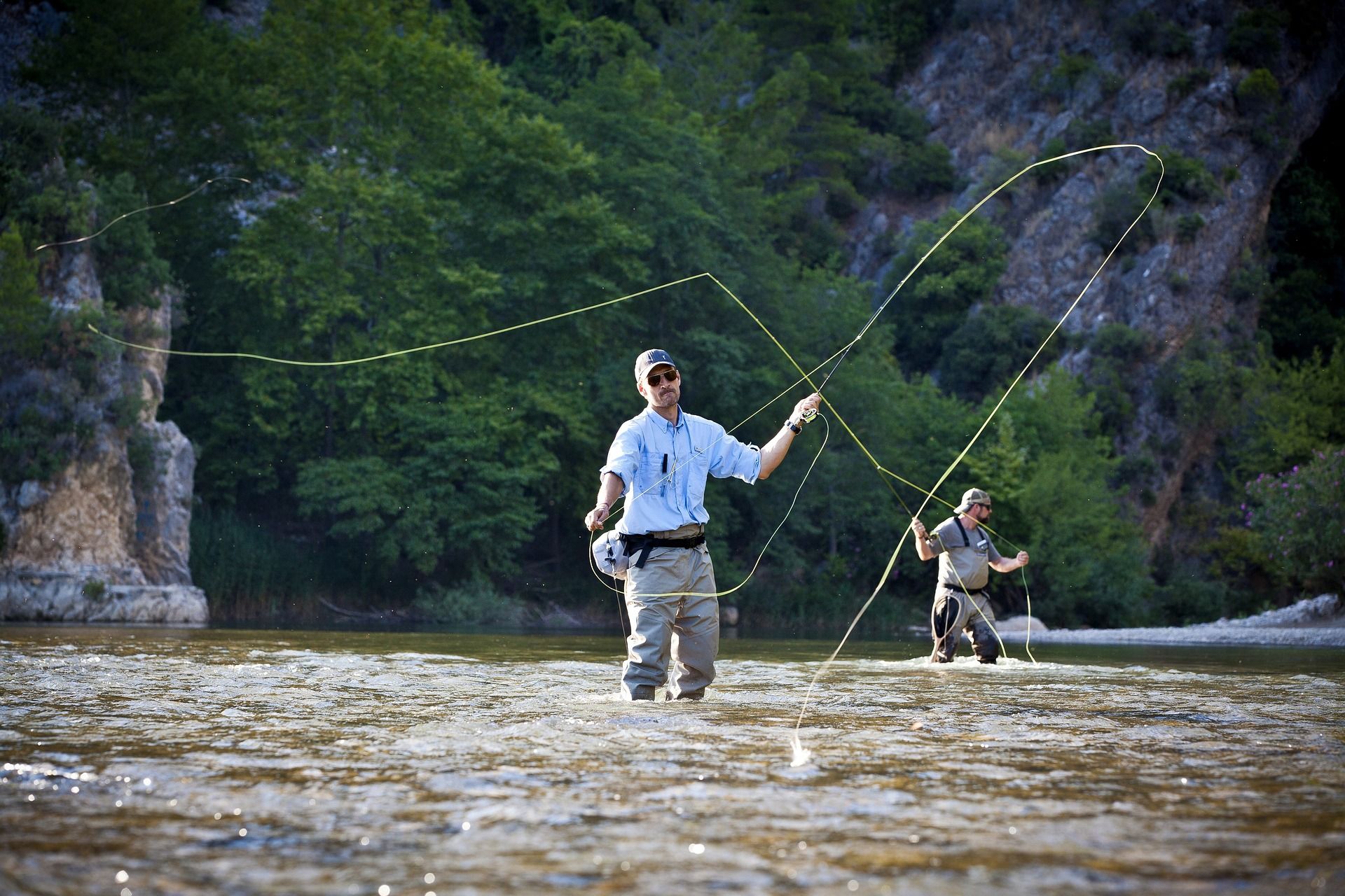 Two men are fishing in a river with trees in the background.