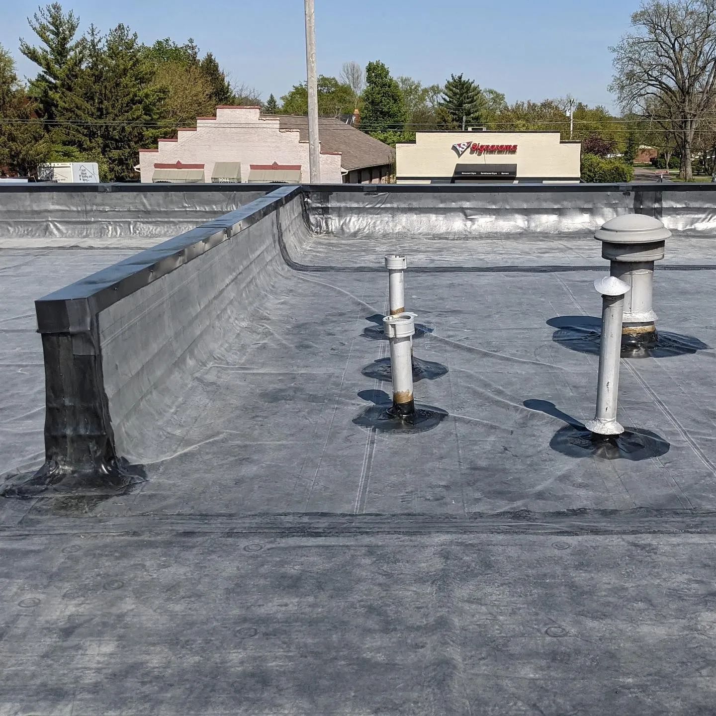 A flat grey roof with a parapet wall and several plumbing vent pipes rising from the surface under a blue sky.