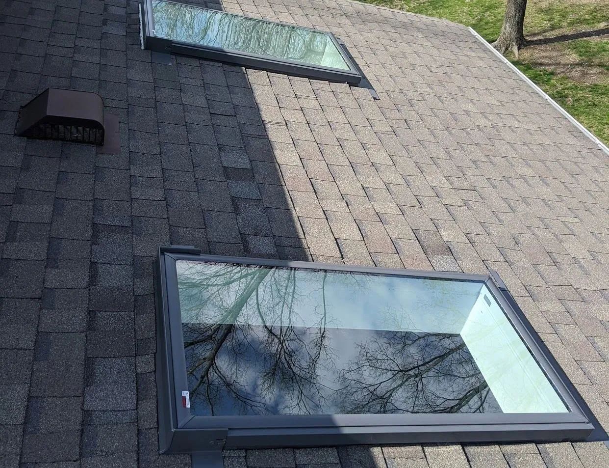 Two rectangular skylights on a gray shingled roof, with a vent nearby. Trees reflected in the glass.