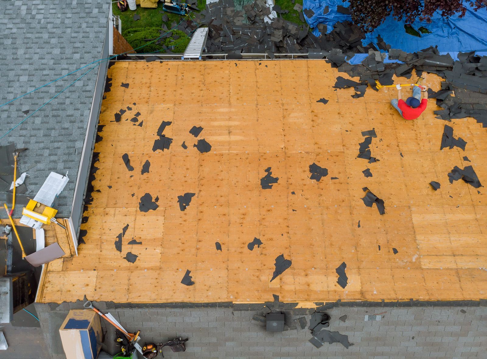 Roofer on a partially torn-off roof, with exposed wood. Green lawn and materials around.