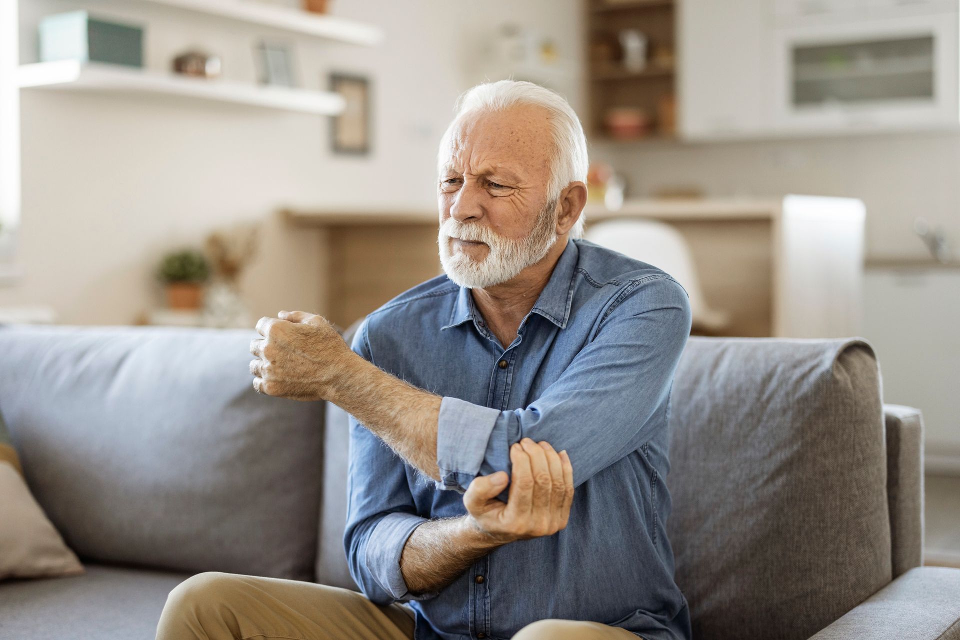 An elderly man is sitting on a couch holding his elbow in pain.