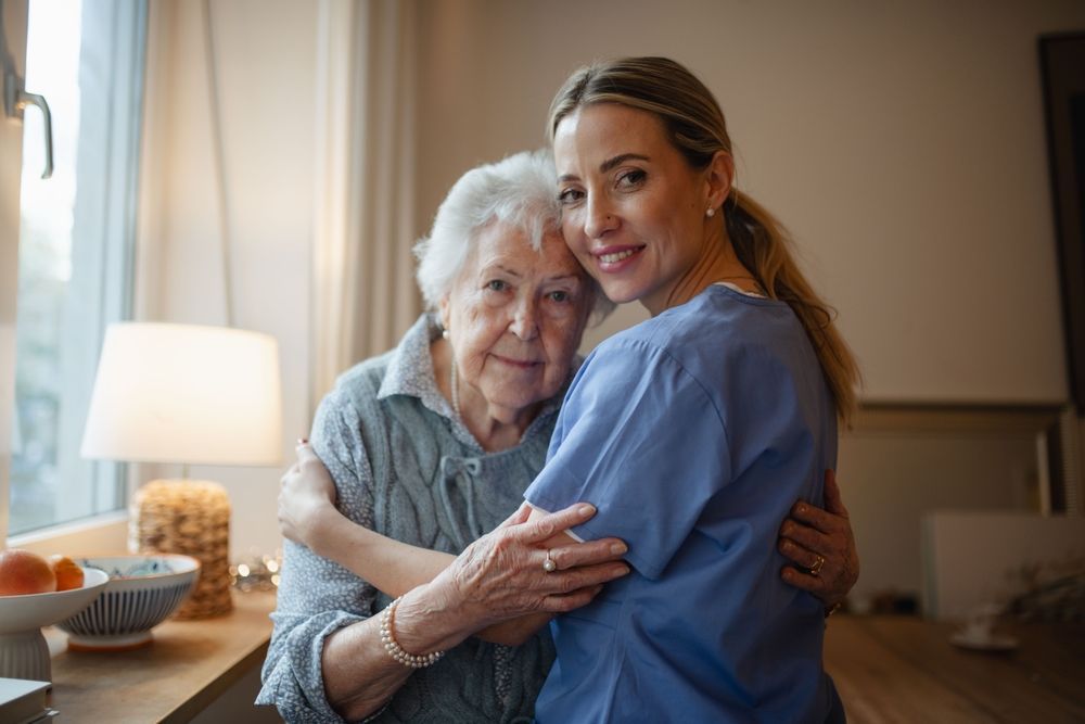 Woman in blue uniform hugs elderly woman, both smiling indoors near window.