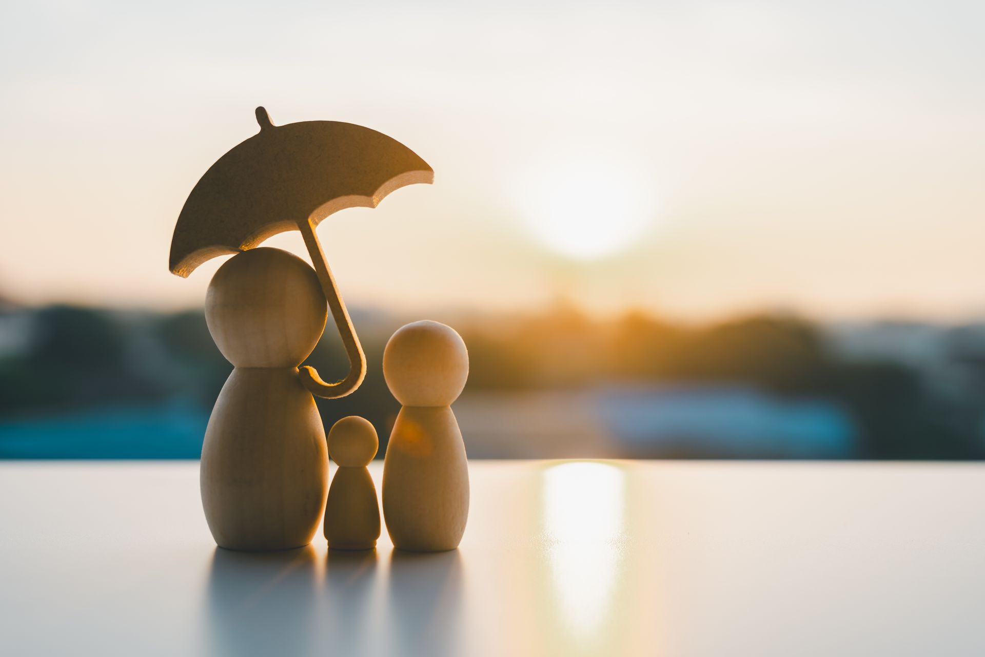 Wooden family figurines under a small umbrella with a sunset backdrop.