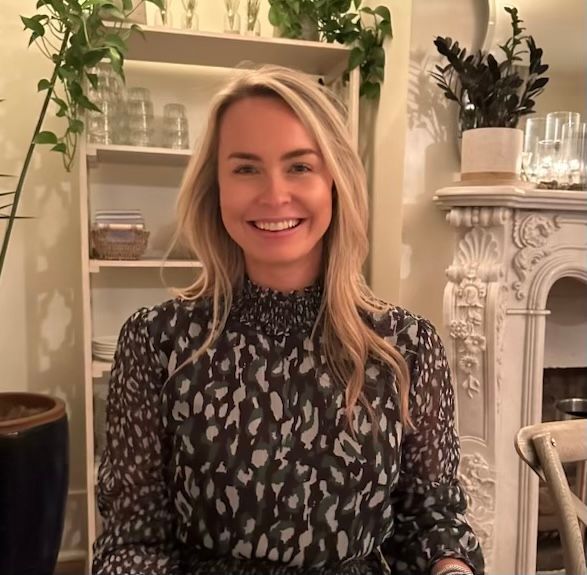 A woman in a blue shirt is smiling in front of a wooden wall.