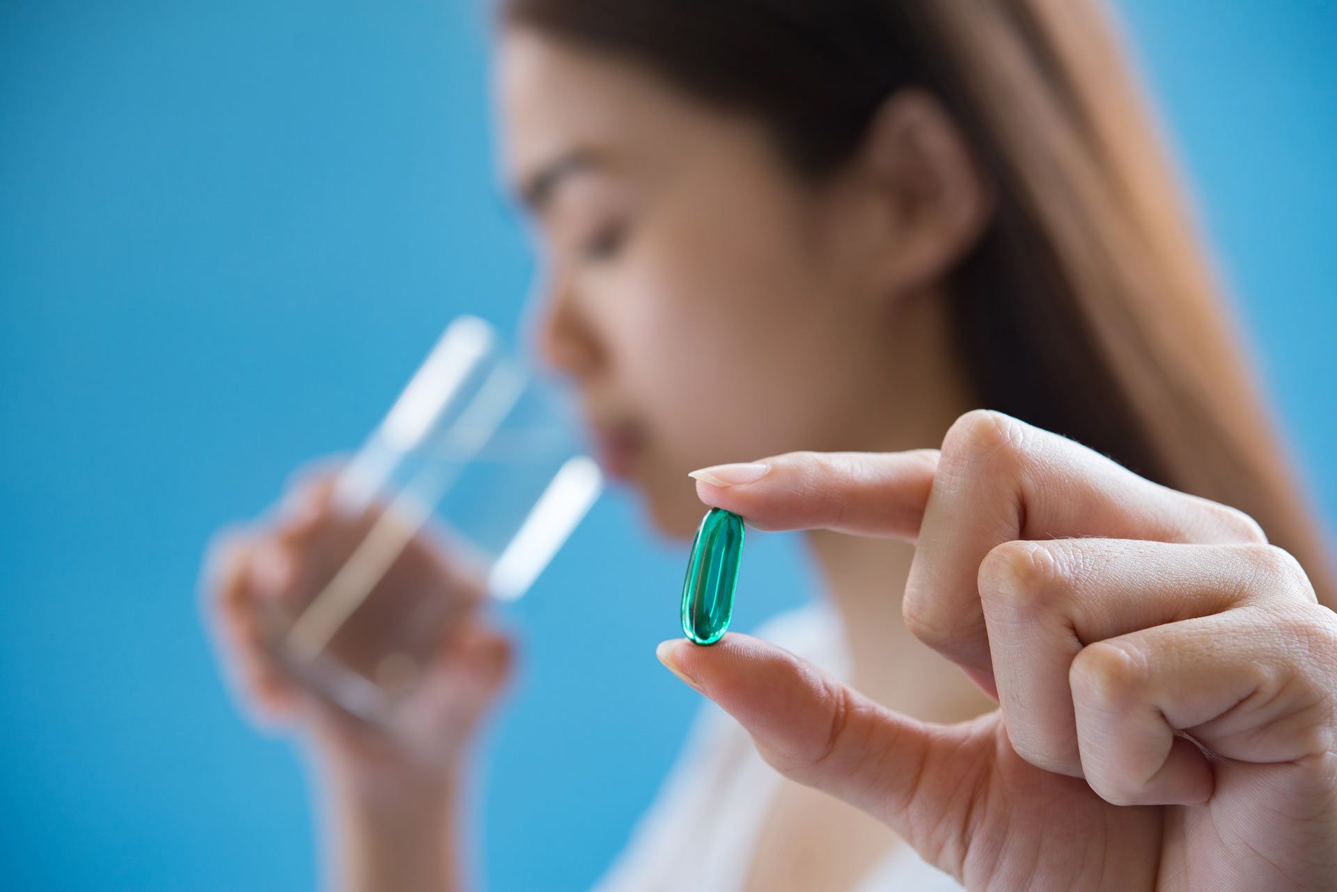 A woman is holding a green pill in her hand while drinking a glass of water.