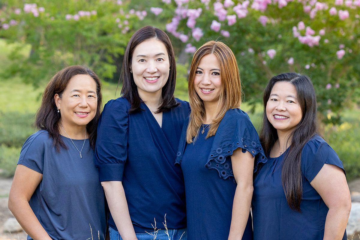 Four people wearing navy blue tops stand side-by-side and smile in a sunny outdoor setting with trees and flowers.