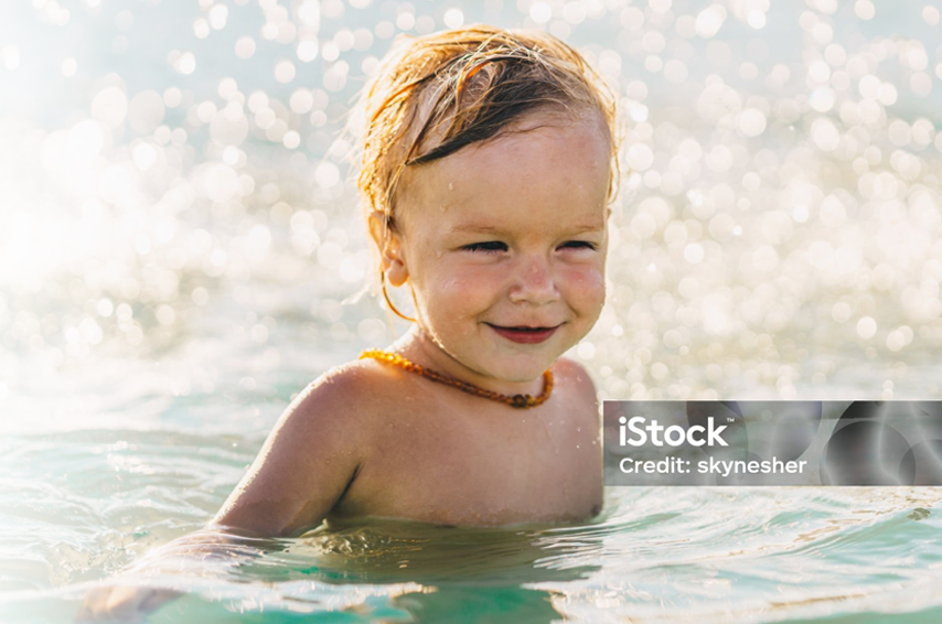 A happy young child wearing an amber necklace smiles while playing in sparkling water with sunlight in the background.