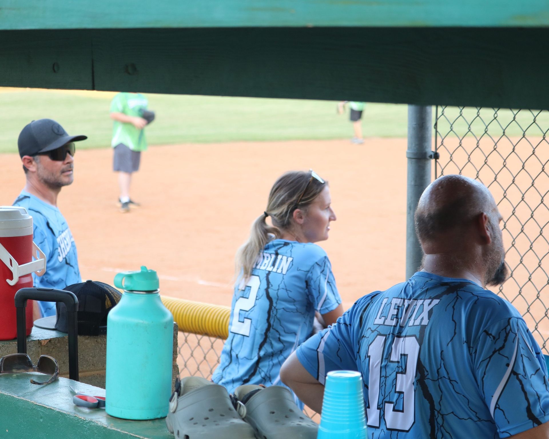 People in softball dugout watching game; light blue jerseys.