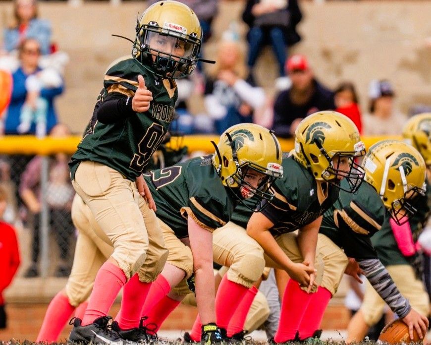 Youth football team in black and gold uniforms posing on a field, holding a trophy and giving thumbs up.
