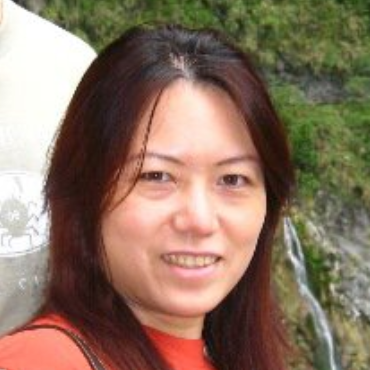 Woman with long auburn hair smiles, wearing an orange top, standing near a waterfall, partially visible person on left.