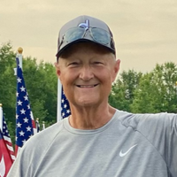 Man in gray shirt and baseball cap smiles, with American flags in the background.