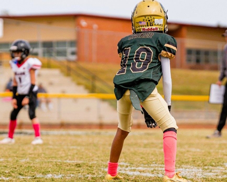 Youth football player in green jersey with number 10, gold helmet, and pink socks, in stance on field.
