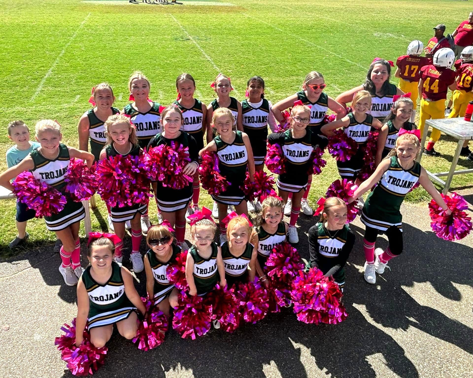 Youth cheerleaders in green and black uniforms holding pink pom-poms on a sunny field.