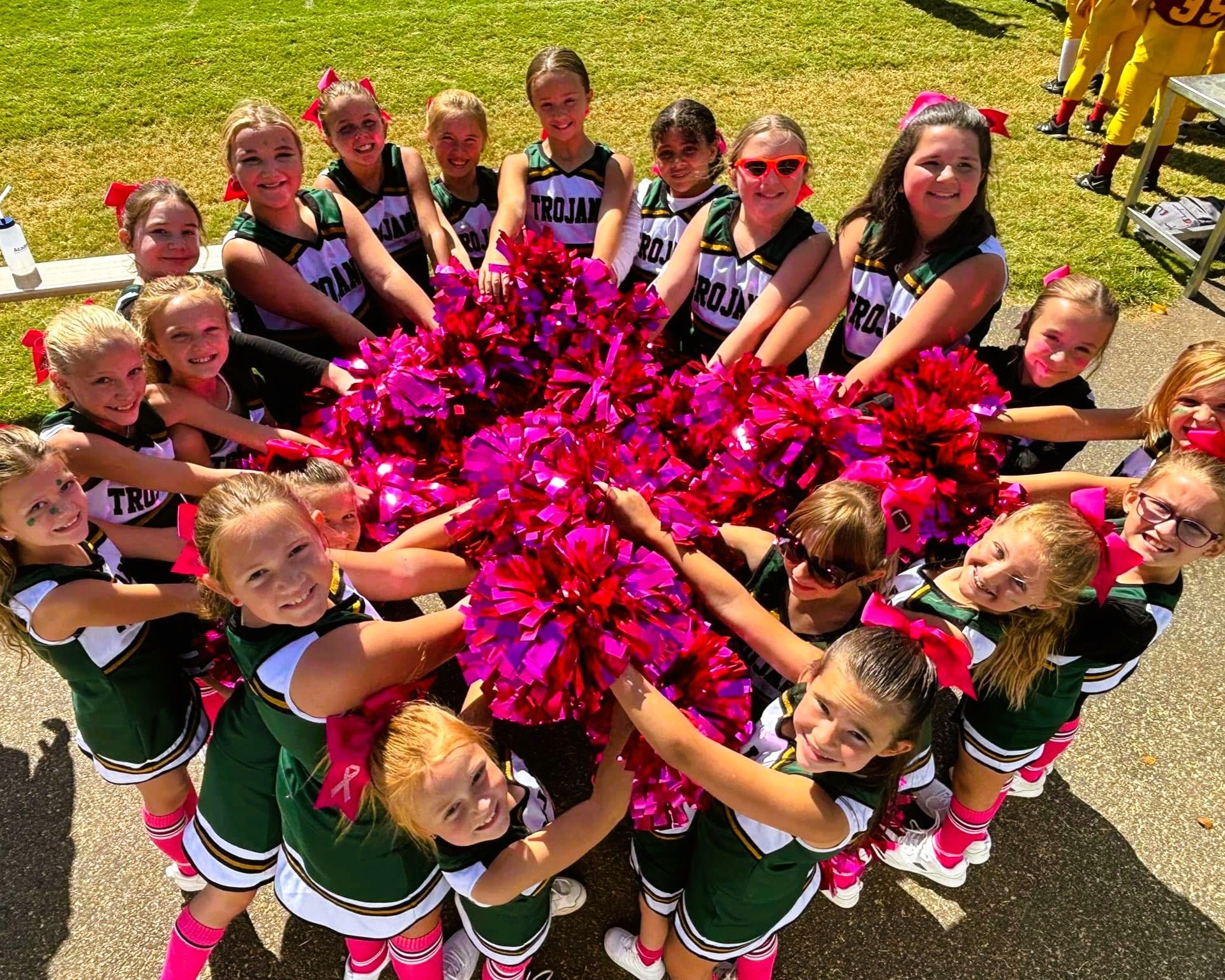 Cheerleaders in green and white uniforms hold up pink and red pom-poms, smiling at the camera.